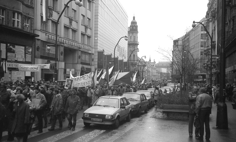 Demonstration in Prag während der Samtenen Revolution im Dezember 1989. Eine Menschenmenge mit Spruchbannern und Fahnen in den tschechischen Nationalfarben läuft durch eine belebte Straße.