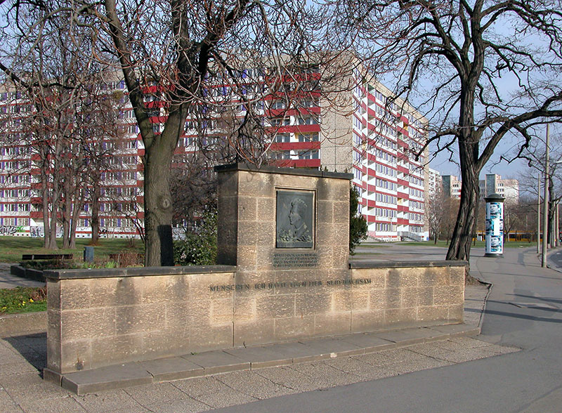 Foto vom Julius Fučík-Denkmal am Straßburger Platz (ehemaliger Fučík-Platz) in Dresden aus dem Jahr 2004. Das Denkmal mit einem Relief von Fučíks Gesicht und der Inschrift „Menschen ich hatte Euch lieb. Seid wachsam“ steht vor einigen Bäumen.