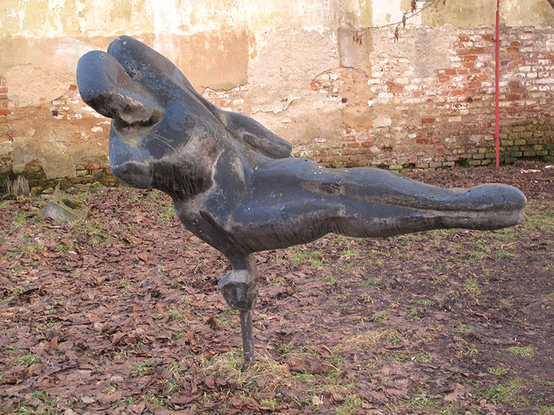 Foto der Skulptur Gymnasta (1979) von Zdeněk Němeček aus dem Jahr 2011. Eine metallene Statue, die zu liegen scheint, ist vor einer Mauer aufgestellt.