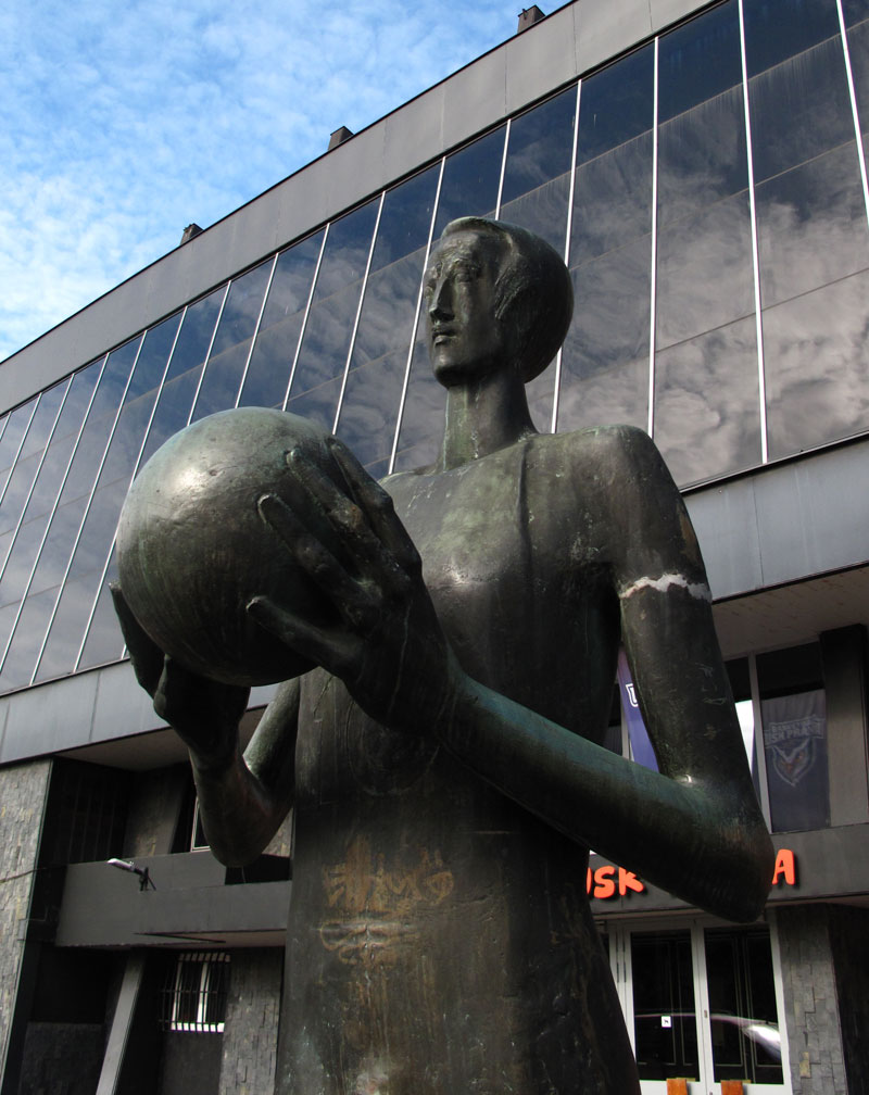 Foto der Skulptur Basketbalista (1977) in Prag von Zdeněk Němeček aus dem Jahr 2019. Eine metallene Statue die einen Basketballspieler mit einem Ball in der Hand darstellt, steht vor einem modernen Gebäude mit Glasfassade.