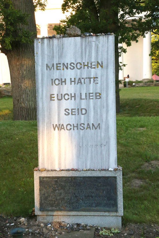 Foto des Gedenksteins für ermordete KZ- Häftlinge in Lauterbach, einem Ortsteil von Putbus auf Rügen aus dem Jahr 2013. Die Stele mit den Worten Menschen ich hatte euch lieb. Seid wachsam steht vor einem Baum. Auf die Stele sind einige Steine gelegt worden.