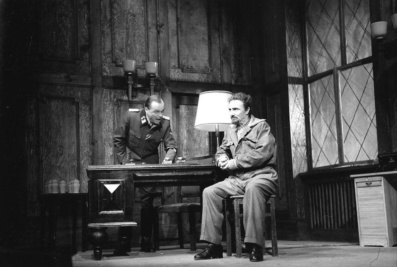 Szenenbild aus dem Stück Julius Fučík von Juri Burjakowski am Deutschen Theater Berlin im Dezember 1951. In der Szene sitzt Fučík (gespielt von Ernst Busch) in einem holzvertäfelten Raum an einem Schreibtisch. Hinter dem Tisch steht ein Beamter der deutschen Besatzung, der Fučík verhört und ihn dabei anschreit.